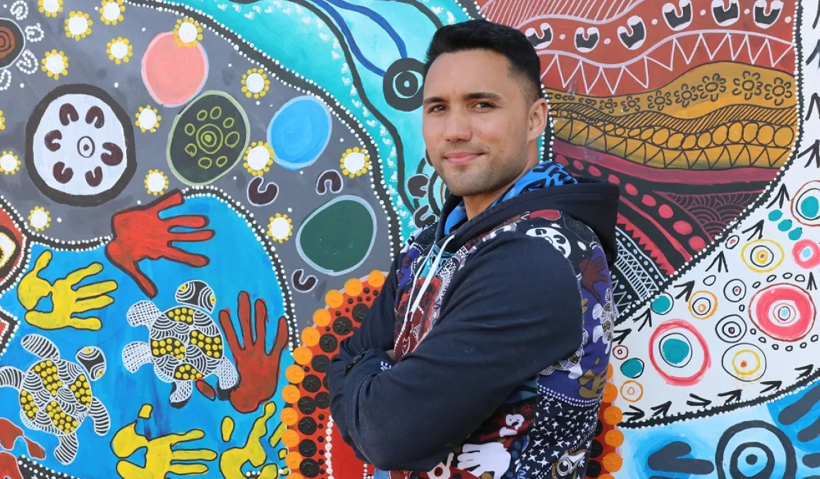 A young adult man in a mainly blue top stands in front of a painting of hands, circles, animals and other features. The painting has blue, grey and red background colours.