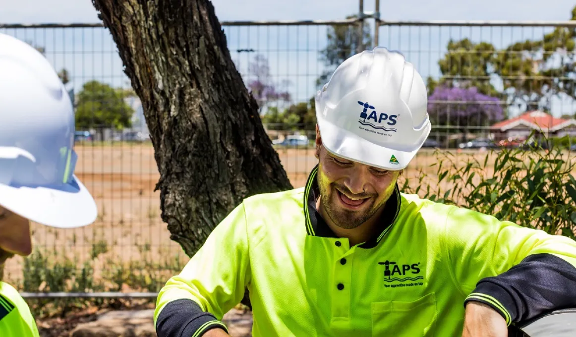 Two men in white hard hats and green work wear tops stand near each other. In the background is a tree, a fence and buildings in the distance.