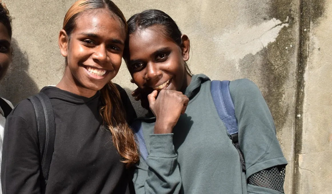 Two Aboriginal young women standing side by side and heads together. The one on the left is wearing a black top and the other an olive coloured top. In the background is a concrete wall.