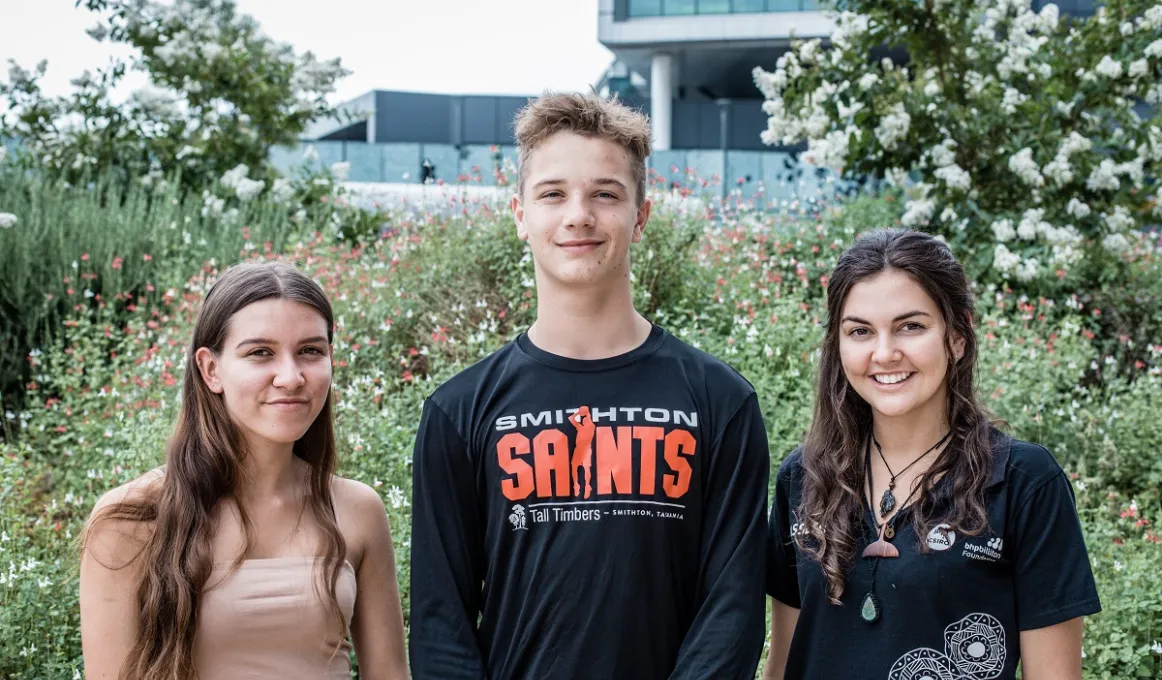 There are 3 young Indigenous people facing the camera and smiling. They are standing shoulder to shoulder, outside on an overcast day, in front of flowering grasses and trees.