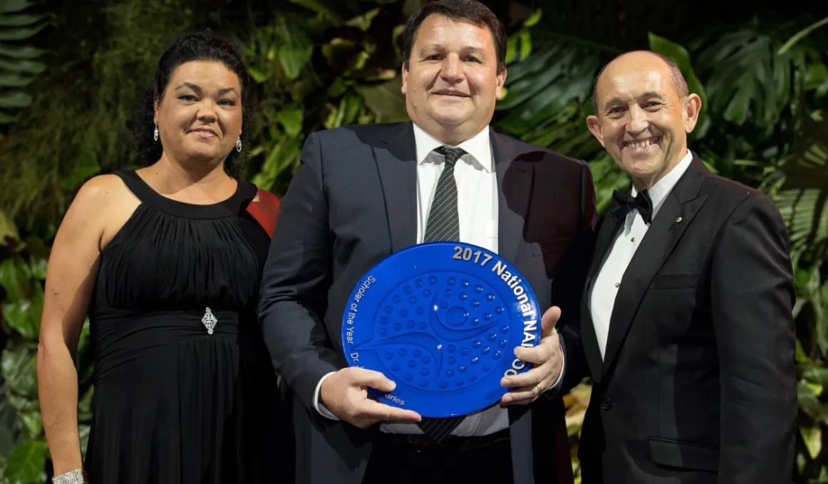 Three people standing together with plants behind them. The man in the middle is holding a large blue plate while a woman is standing to his right and man to his left.