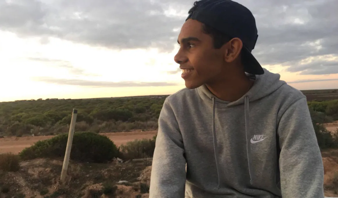 Young Aboriginal man wearing a grey hoody and cap looks out over bush landscape with cloudy sky in the background.