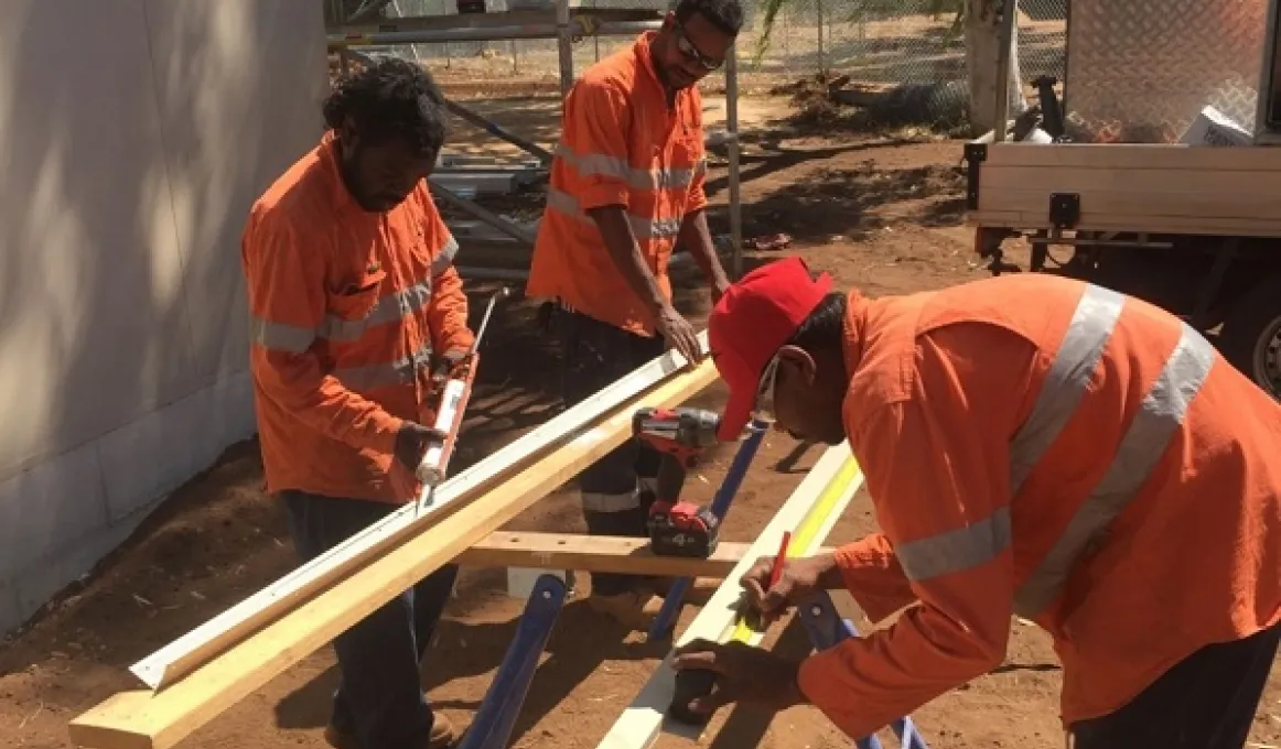 Three Aboriginal men dressed in orange work clothing and standing on red dusty soil next to a building prepare timber for use in construction.