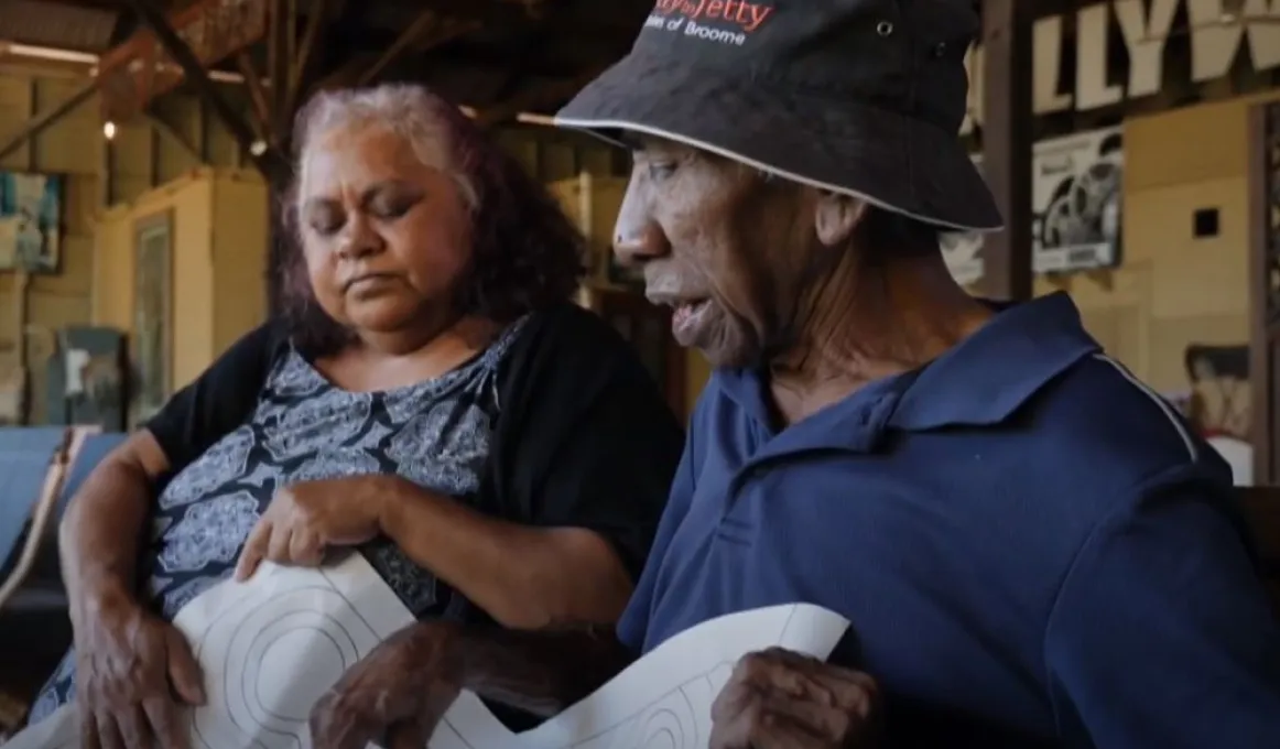 An Aboriginal woman in dress and dark cardigan sits next to an Aboriginal man in hat and blue shirt. In the background is a wall with posters and paintings on it.