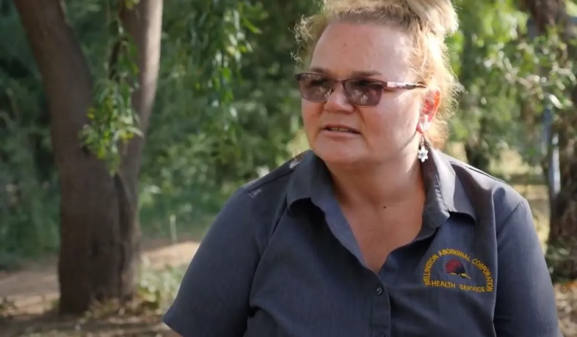 A head and shoulders shot of a woman with her hair pulled back, wearing glasses and a grey shirt. In the background are trees.