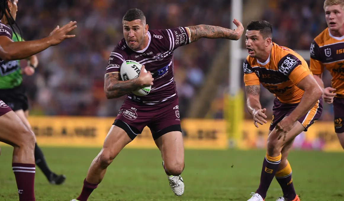 Indigenous man in maroon sports uniform and holding a ball runs past a man in a yellow uniform. At left and right are more men and in the background is grass, goals posts and a crowd of people.