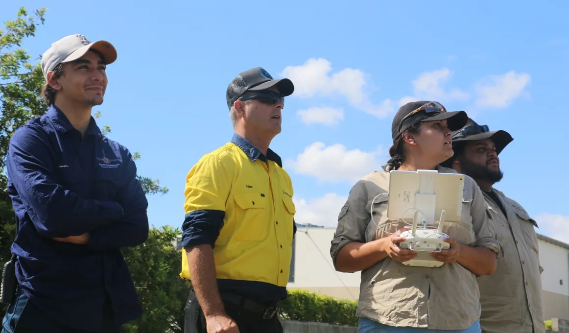 Three men and one woman dressed in work wear or casual wear stand shoulder to shoulder. The woman holds a white drone controller. All are looking up into the sky.