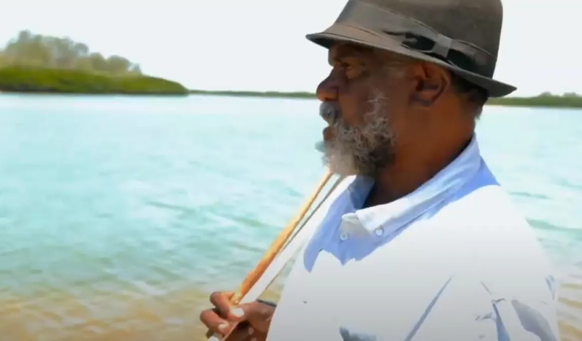 An Indigenous man in hat and pale shirt holds a spear over his shoulder. In the background is water and foliage.