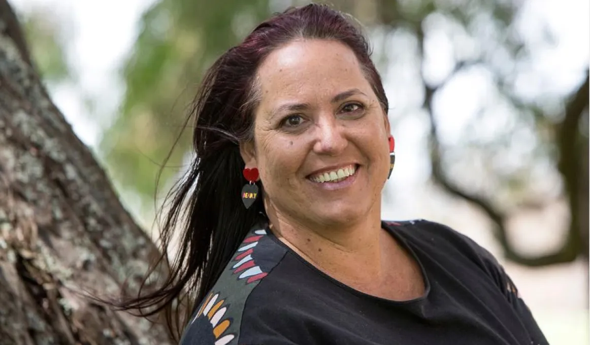 A middle-aged woman with long dark hair leans against a tree. She wears a dark top with colourful patterns around the shoulder area. In the background are more trees.