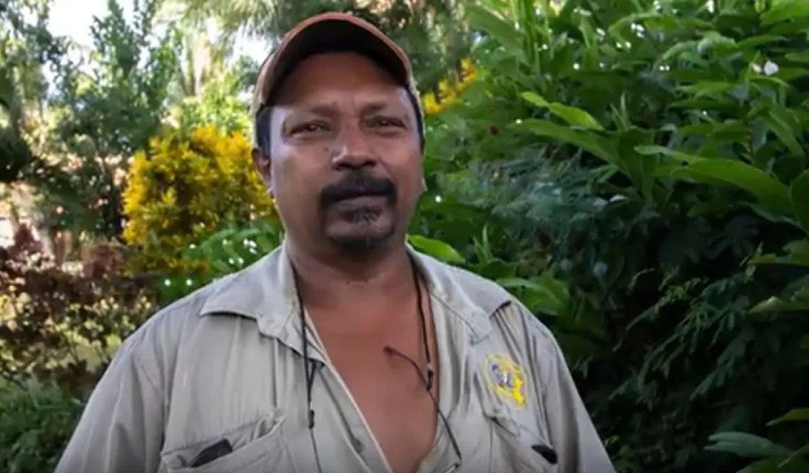 An Indigenous man in khaki shirt and wearing a cap stands in front of green and yellow and brown foliage.