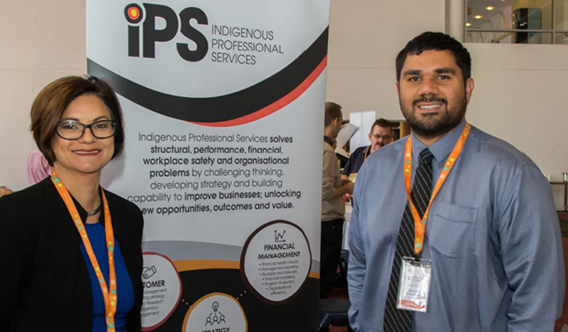 Indigenous woman in black jacket stands with Indigenous man in blue-grey shirt in a room with other people. Between them is a sign with logos and words including IPS Indigenous Professional Services.