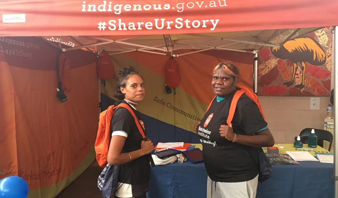 Two young Indigenous women wearing orange backpacks and dressed in Batchelor Institute shirts stand in front of a blue table topped with papers and pamphlets set up in a stall displaying the words: indigenous.gov.au #ShareUrStory.