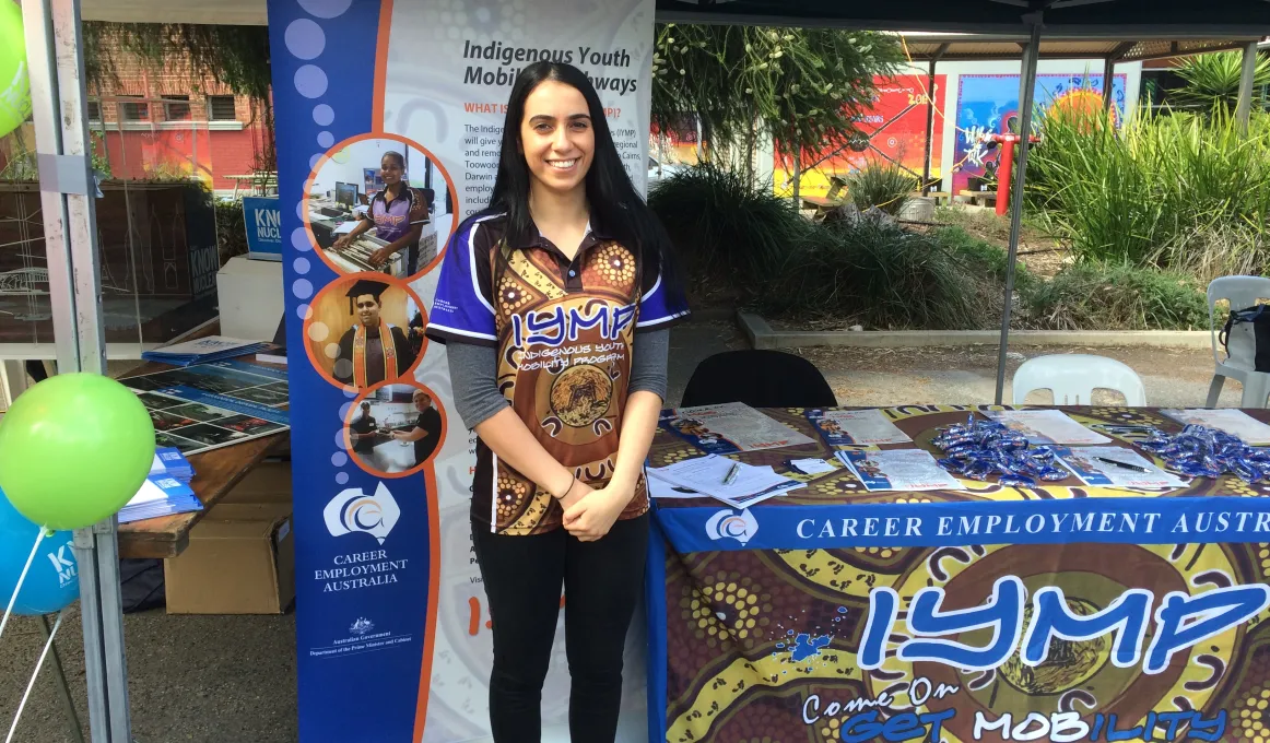 Young Indigenous woman standing in front of sign and to the side of a display table for Indigenous Youth Mobility Pathways. She is dressed in black pants and a colourful shirt showing Indigenous designs.