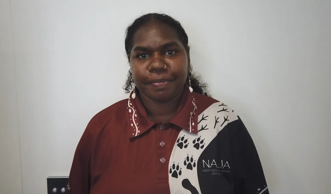 An Aboriginal woman with hair tied back wears an ochre red, white and black shirt with Aboriginal designs over the left shoulder. In the background is a white wall.