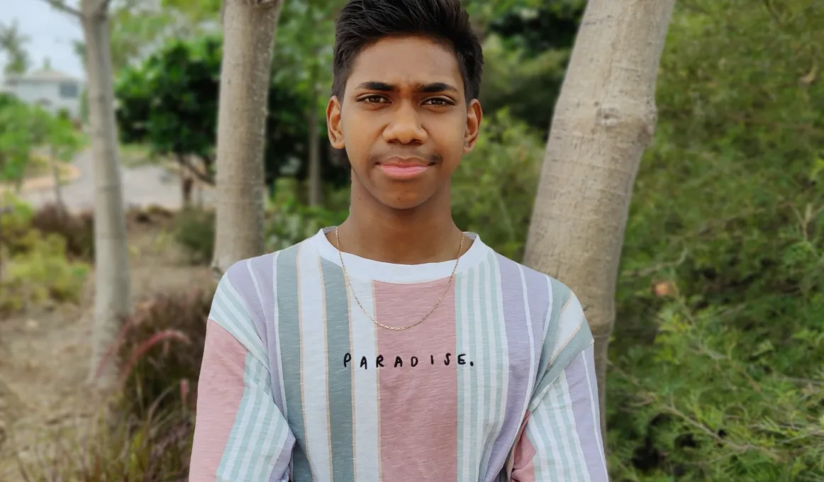 A young man in a colourful shirt with vertical stripes stands in front of some trees beyond which ae bushes and a road.