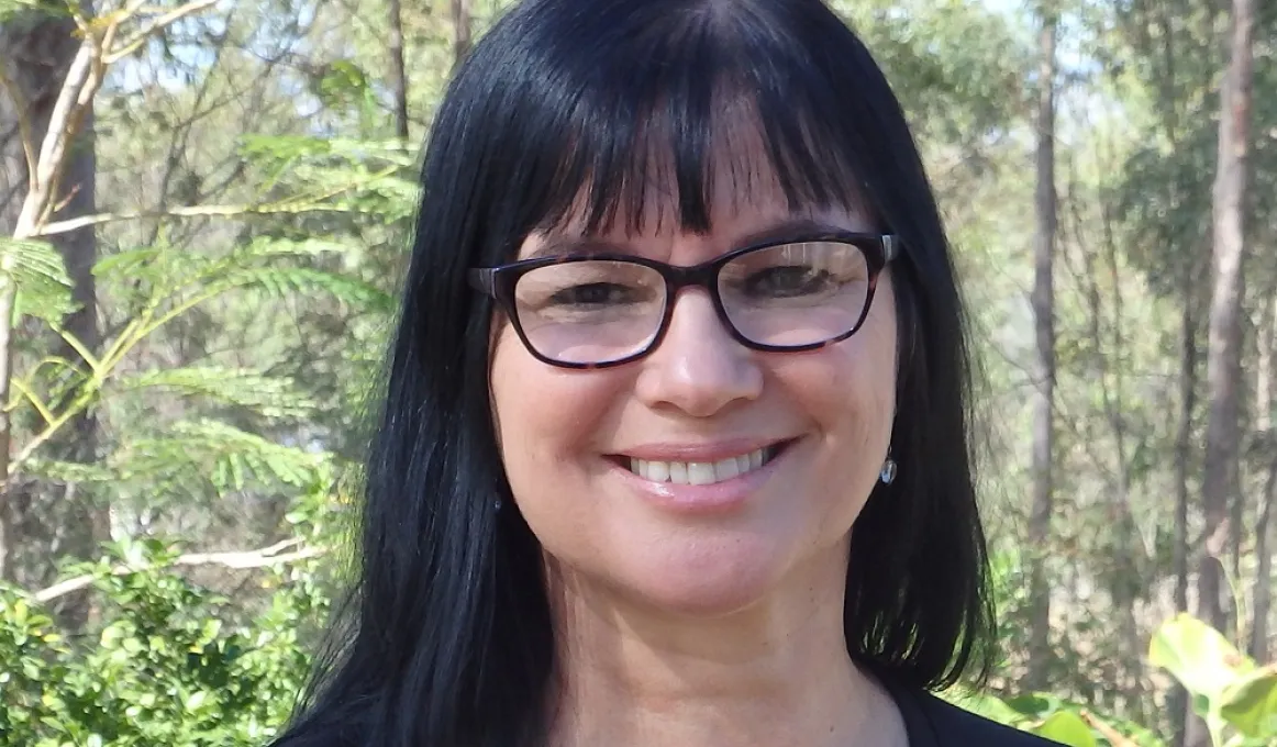 A woman with long black hair and wearing glasses faces the camera and smiles. In the background are trees and shrubs.