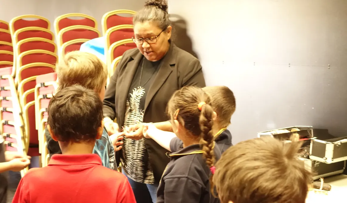 An Indigenous woman in brown coat stands with five children in a room with stacked chairs in the background and a table with craft making items.