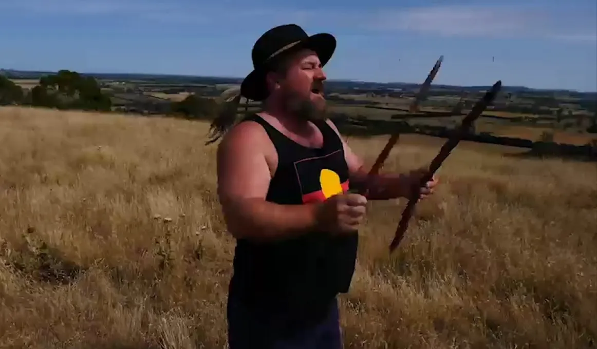 An Aboriginal man in black hat and black singlet with Aboriginal flag on it holds two sticks as he stands on a hill covered in dry grass. In the background is a blue sky, more hills and fields and trees.