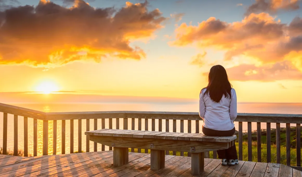 A woman with long dark hair and wearing a light jacket and dark pants sits on a bench overlooking water. In the background is a darkening sky, clouds and a setting sun.