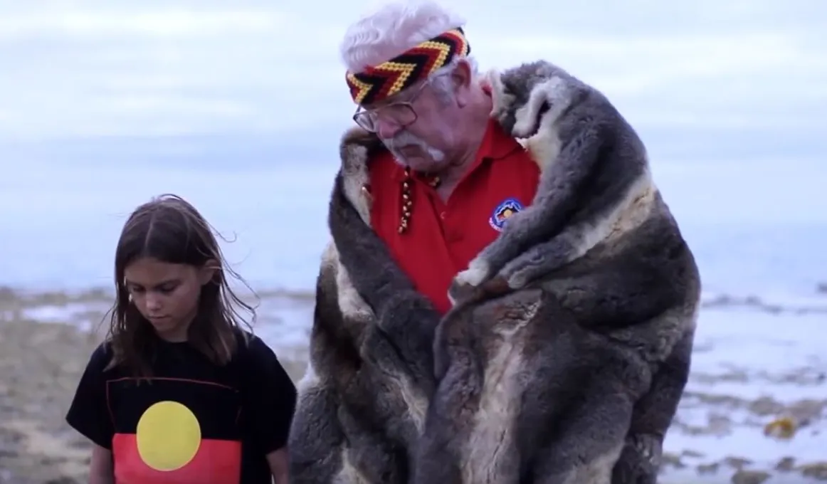 A young girl dressed in a t-shirt with Aboriginal flag design (black bar at top, red at bottom and yellow disc in middle) stands with an elderly man dressed in red shirt and fur cloak in front of a seascape.