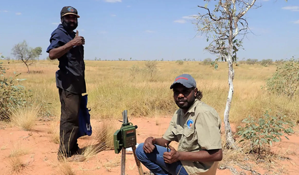 Aboriginal ranger in blue shirt stands and shows the thumbs up sign. Before him an Aboriginal ranger squats on sandy soil in front of a camera on a post. In the background are trees, grass and blue sky.