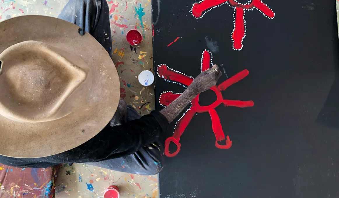 A man in a brown hat and dark shirt sits next to a black canvas on the floor. He is painting red starfish like figures.