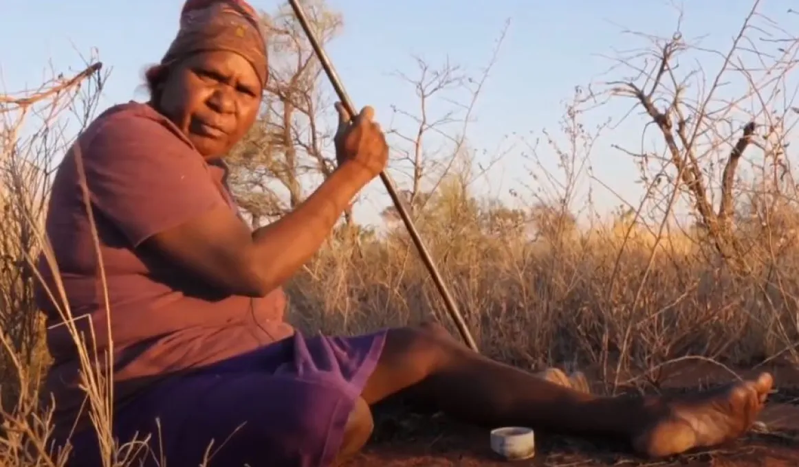 A mature Aboriginal woman dressed in top and skirt sits on red soil in amongst long grass. She holds a metal rod in her right hand.