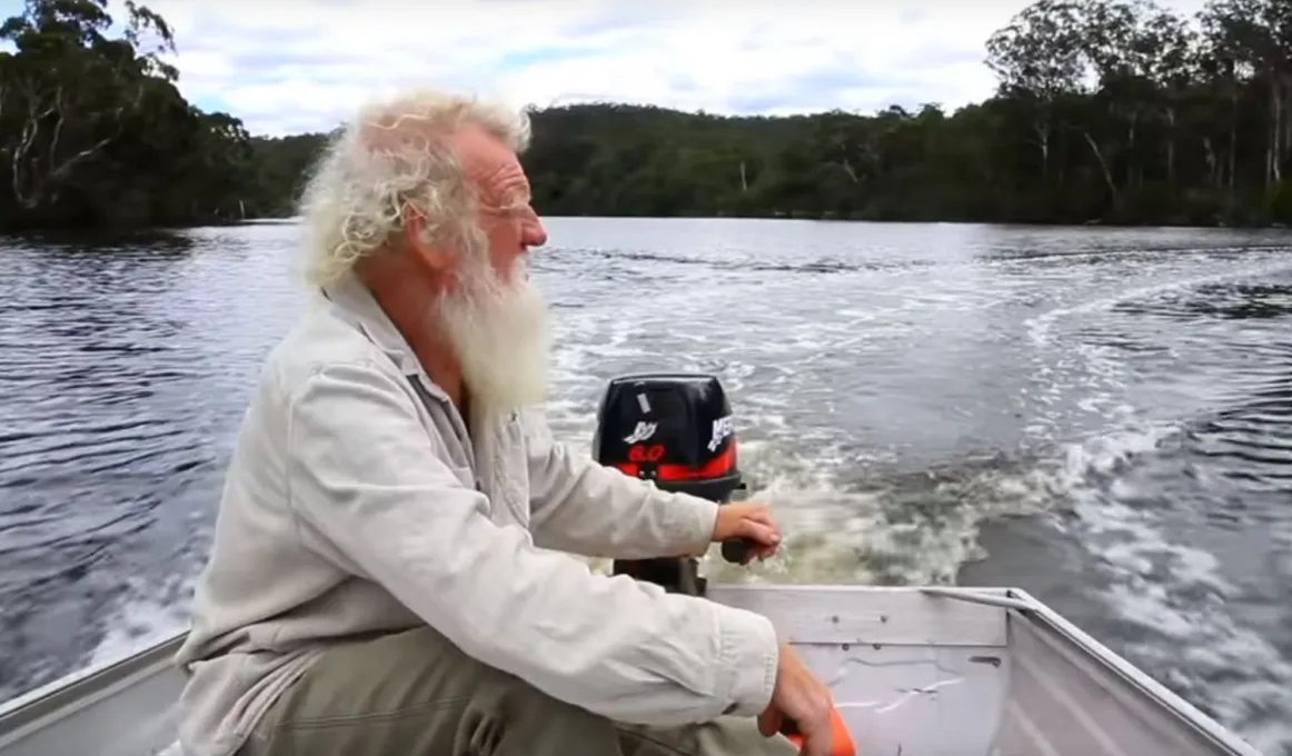 A white haired and white bearded man wearing a pale shirt sits in a boat with an outboard motor on a large river with trees in the background.