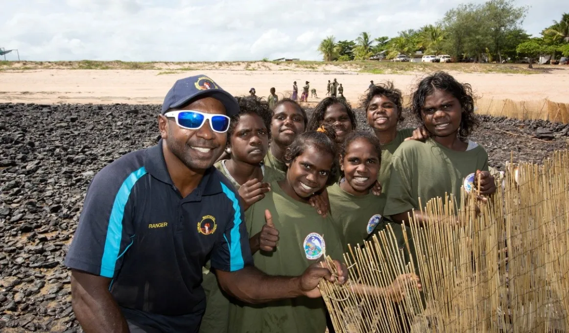 Aboriginal man in blue shirt and cap with Aboriginal youth in green shirts stand on rocks at a beach with sand and trees in the background. He supports an upright bamboo barrier.