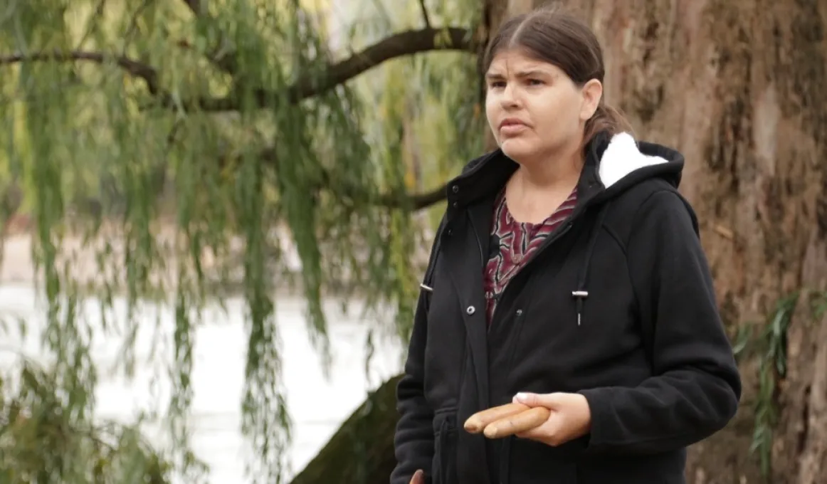 A woman in black top stands in front of a tree. In the background is a river and beyond that a riverbank and foliage.