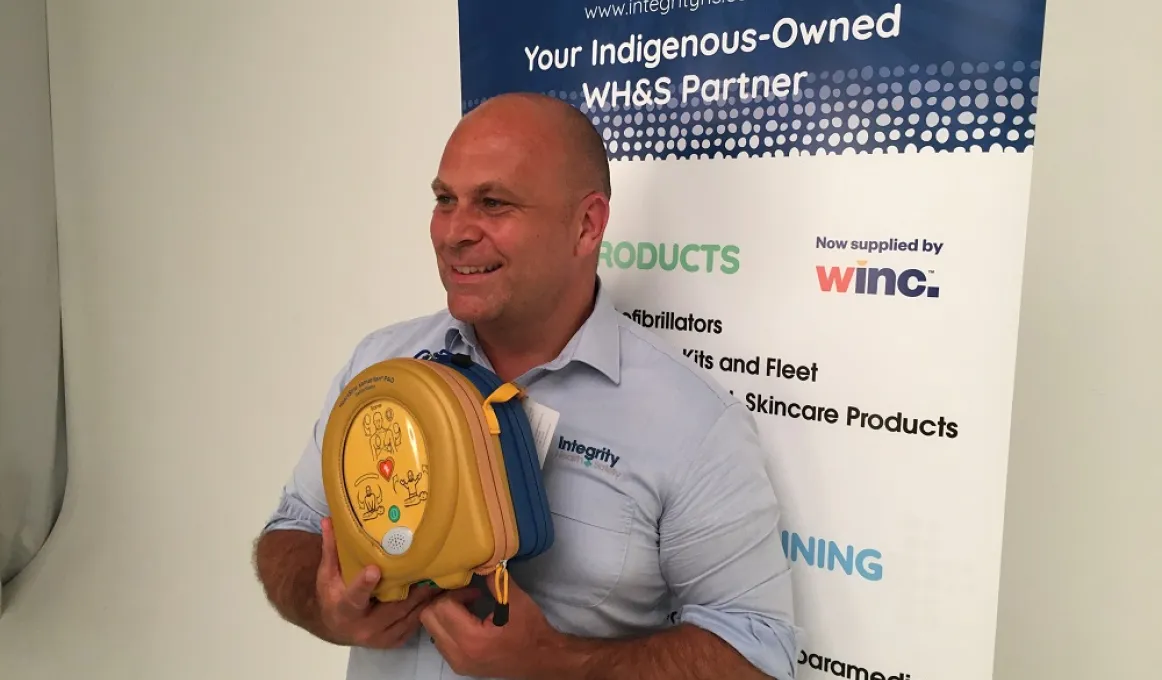 A man in a blue shirt holds a defibrillator as he stands in front of a poster which features the text: Integrity Health & Safety. Your Indigenous owned WH&S Partner.