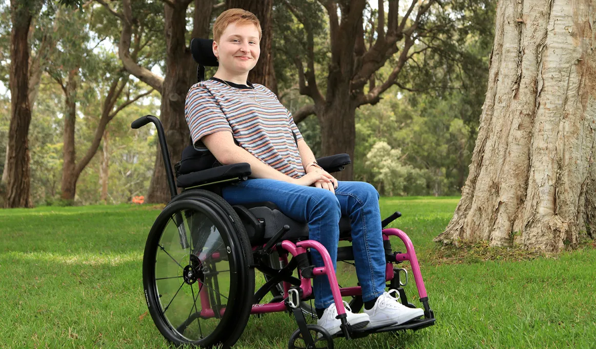 A young woman sits in a wheelchair on grass. In the background are trees.