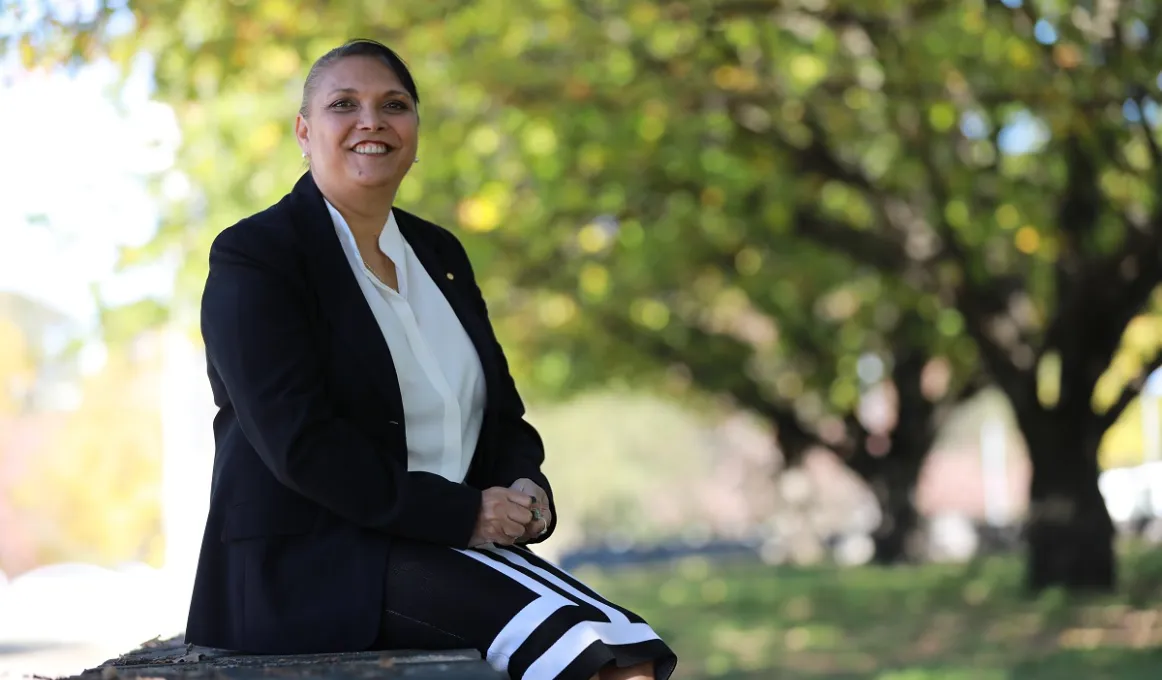 Indigenous woman in black jacket and white blouse sits in a wall in a park. In the background are trees and grass.