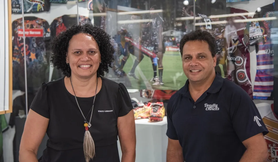 An Indigenous woman dressed in black and an Indigenous man in a dark shirt look to camera. In the background is a display of shirts, boots and posters.