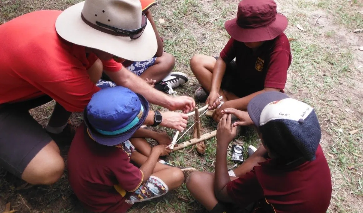 Man in hat teaches four Indigenous students in hats and maroon shirts how to start a fire