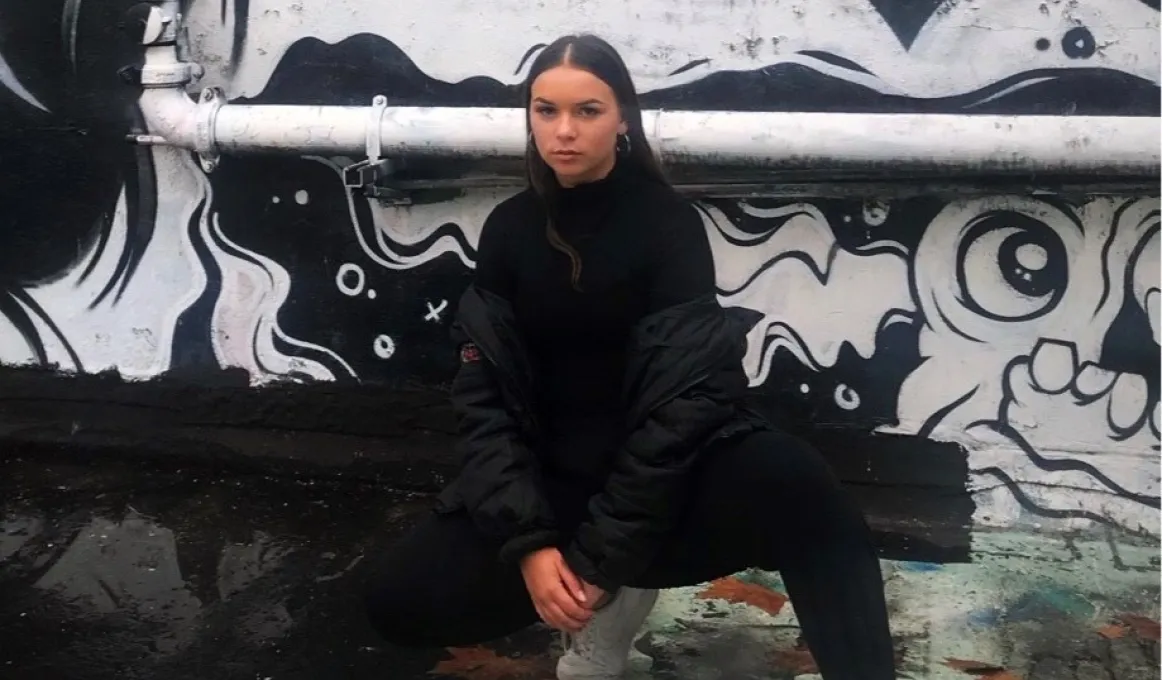 A young woman with long brown hair dressed in dark top and pants, and white shoes squats in front of a wall painted with black and white images. A pipe runs horizontally across the wall.