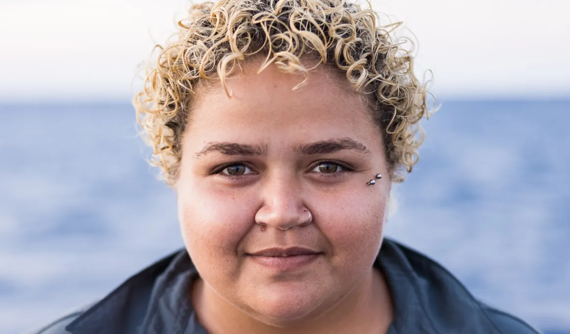 Head and shoulders shot of a young women with blonde, curly hair wearing a blue grey shirt.