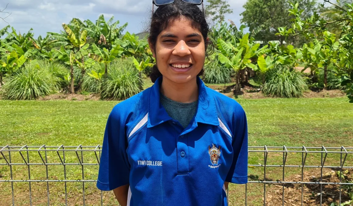 Young woman in blue shirt stands in front of a metal fence. In the background is grass and trees and a cloudy sky