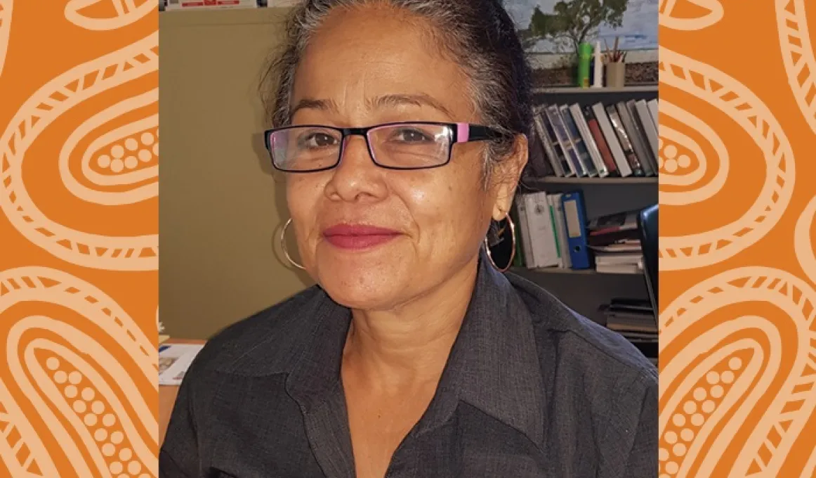 Indigenous woman with hair drawn back and wearing glasses and a dark shirt. Behind her is shelving and other office content.