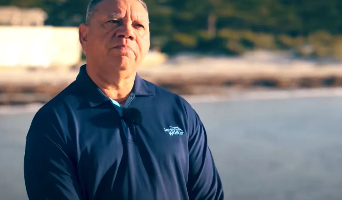 Middle-aged man in blue top stands with water behind him. Also in the background is a shoreline and trees.