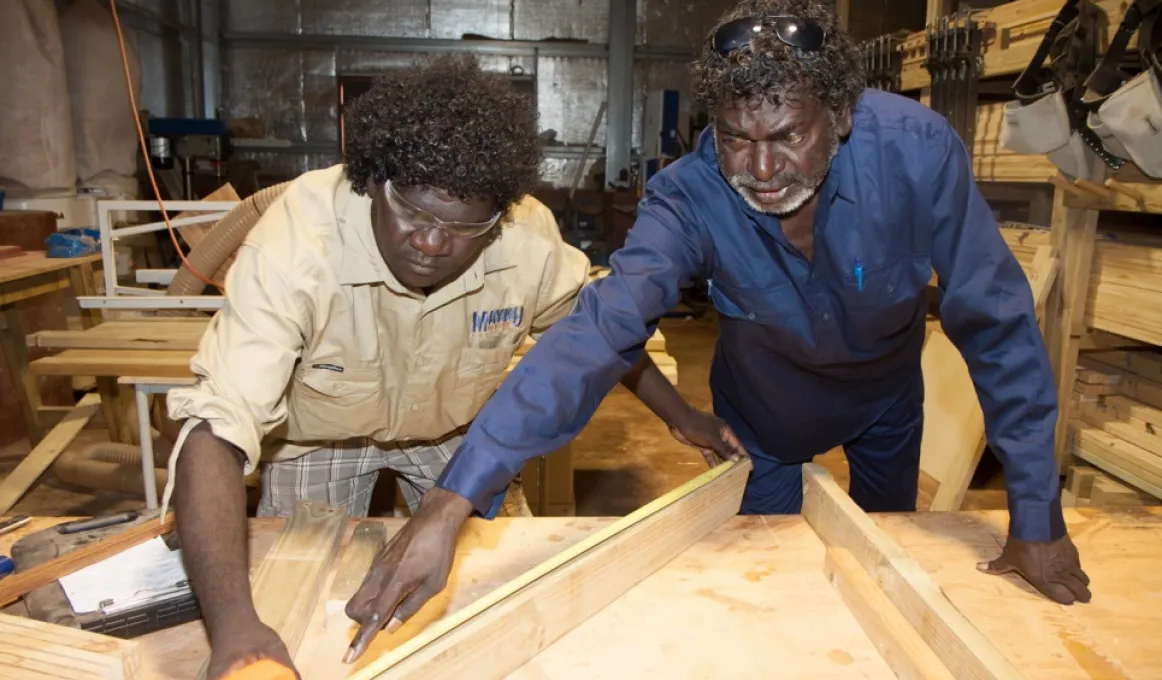 Two Indigenous men working at a wood working bench with the elder man directing the younger