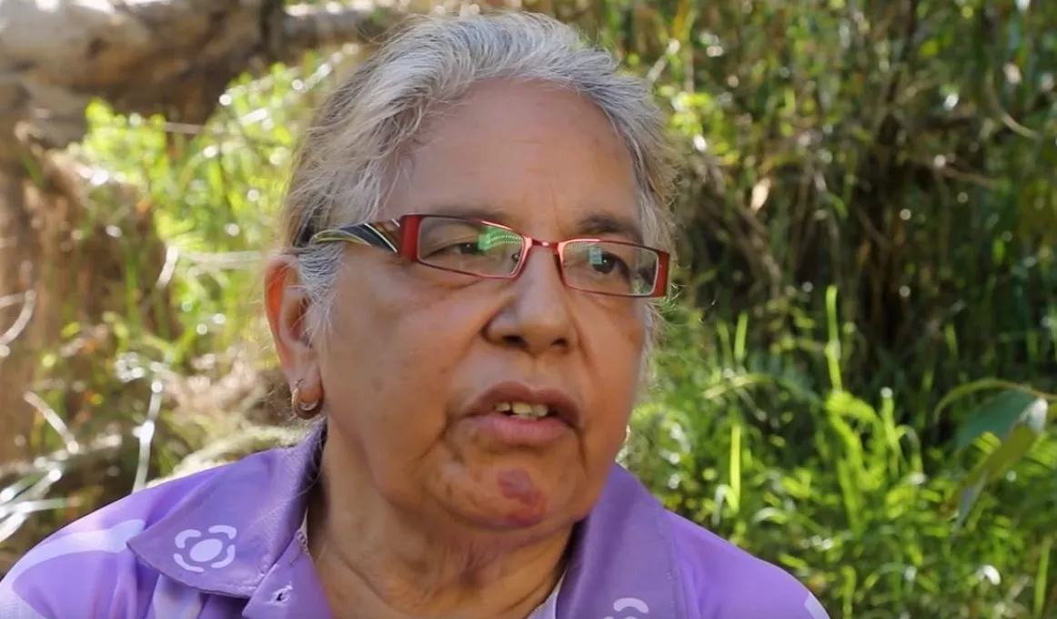 Head and shoulders photo of an elderly woman with greying hair and wearing glasses and a purple shirt. Behind her is thick green vegetation.