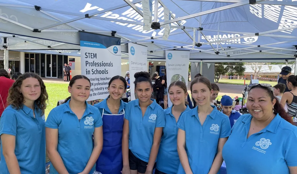 Six young women and one adult woman dressed in blue tops face the camera. In the background are more people and displays. Above them is the framework and roof of a large gazebo.