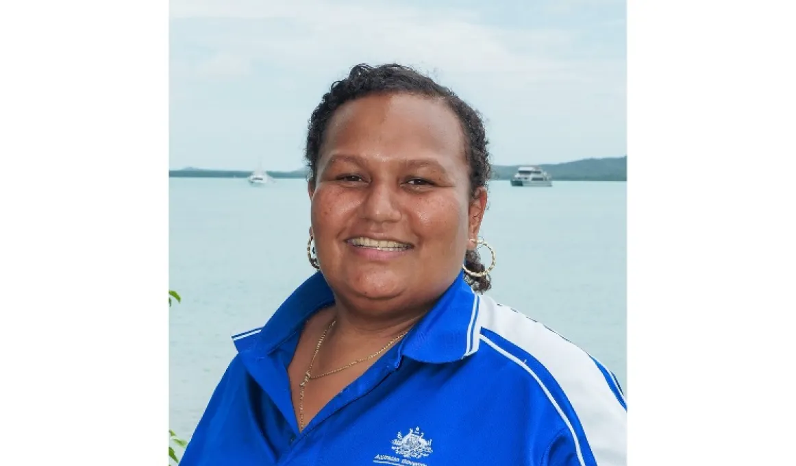 A woman in blue and white polo shirt faces the camera. In the background is blue water, two boats and some land.