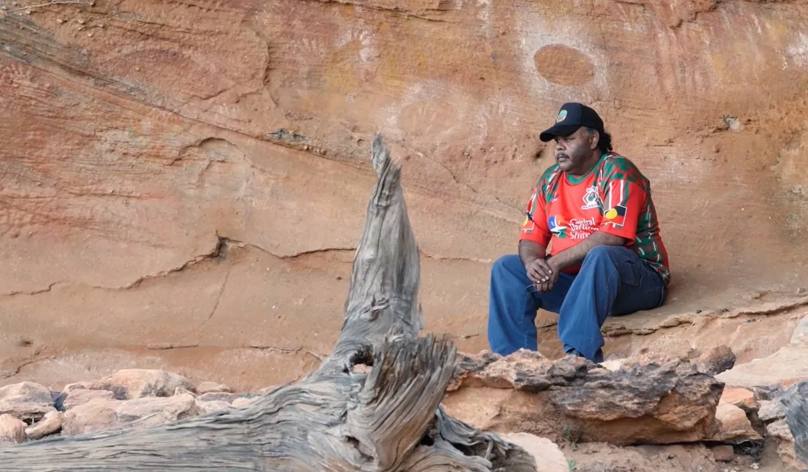 Aboriginal man in red shirt, blue jeans and a black cap sits on a rock ledge behind which are cave paintings. In front of him is a fallen log.