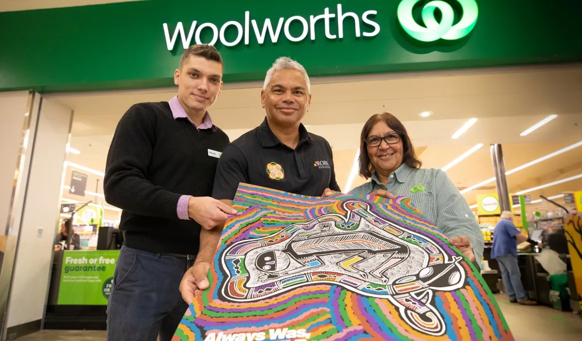 Three people hold a poster featuring an image of the Australian landmass, in front a Woolworths store. Above is the Woolworths name and logo. In the background is the store with shoppers.