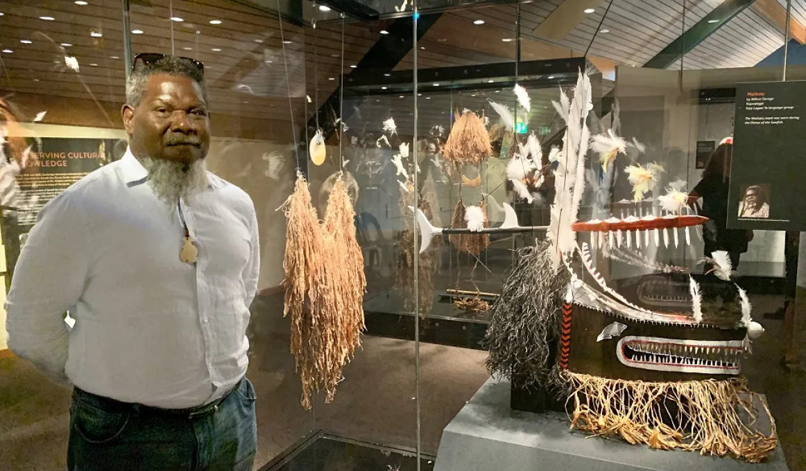 Indigenous man in pale shirt and jeans with a beard, stands next to a yellow mask of draping filaments suspended on wires from above. In the background and panes of glass and more Indigenous masks and items.