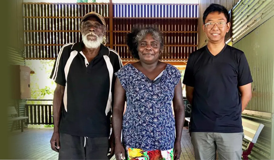 Three people stand on a wooden floor and behind them is a covered area with walls and ceiling but opened to the outside. An Aboriginal man in dark shirt and shorts is at left. Next to him is an Aboriginal woman in blue shirt and colourful skirt. At her le