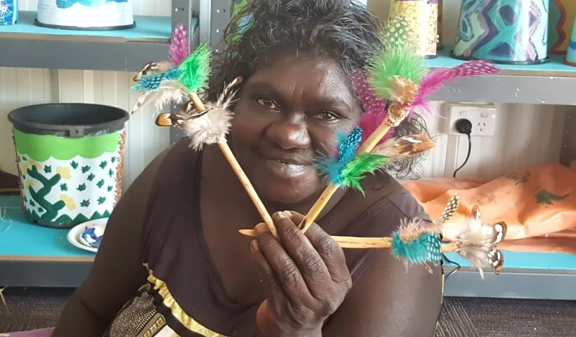 An Indigenous woman dressed in brown dress holds three items of jewellery made from feathers and sticks. In the background are shelves holding painted tins.