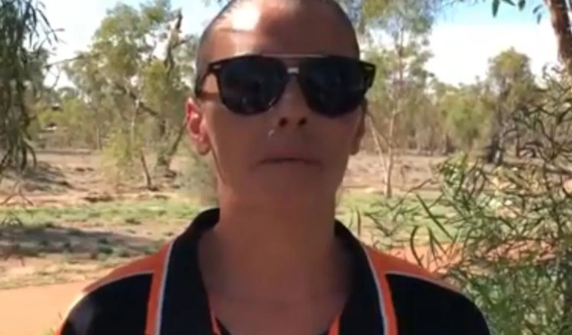 Woman wearing sunglasses and black and orange shirt stands under a tree with bushland in the background.